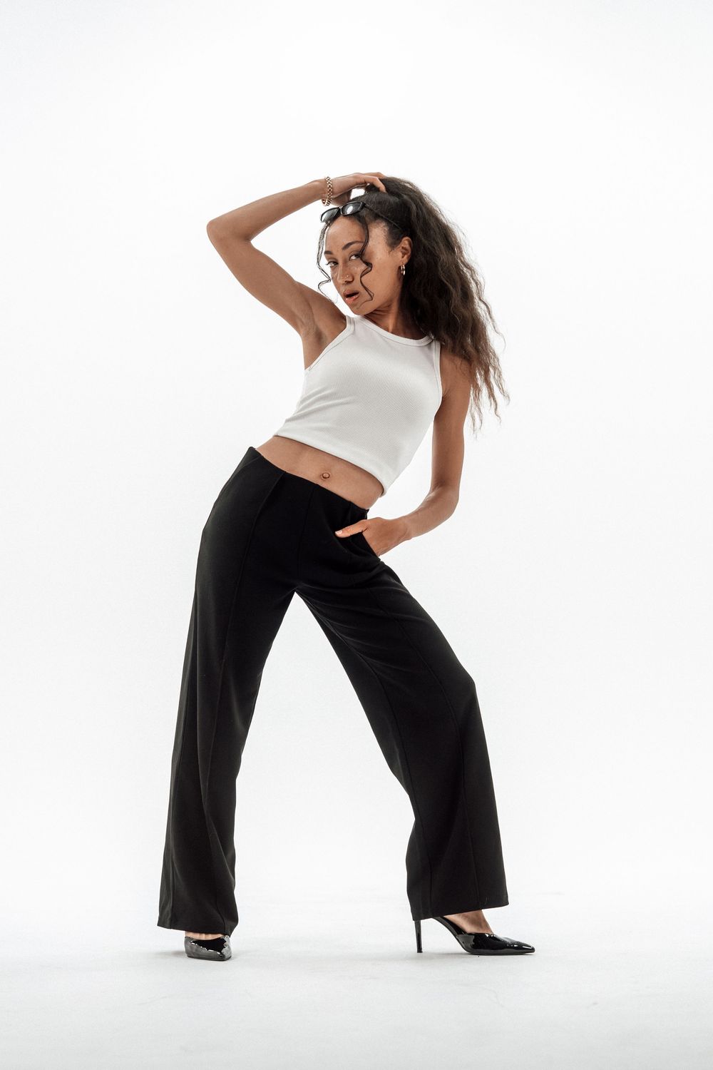 Woman posing in white crop top, black wide-leg pants, and black heels against white background.