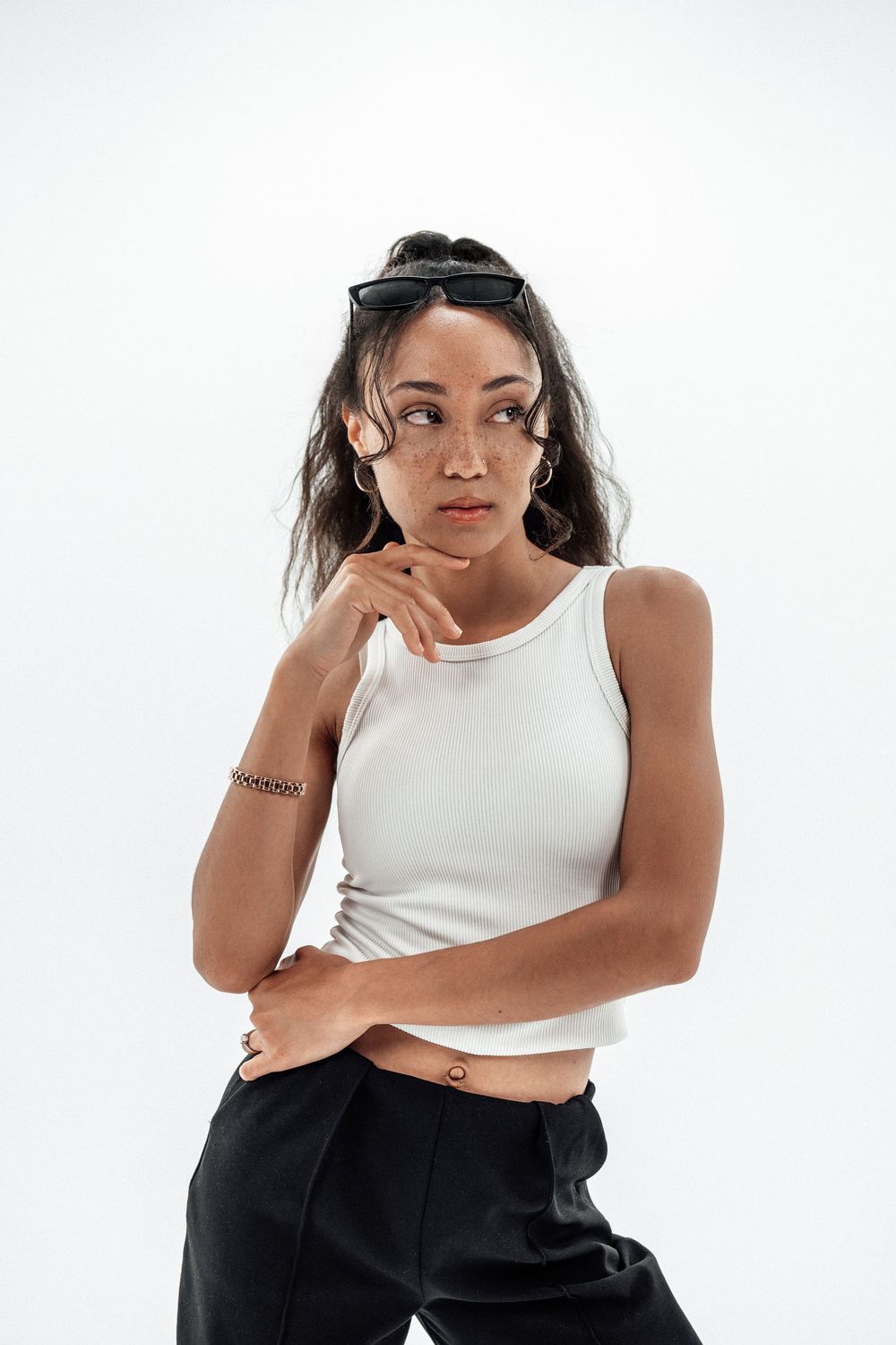 Young woman in white crop top and black pants poses confidently with sunglasses on head against white background.