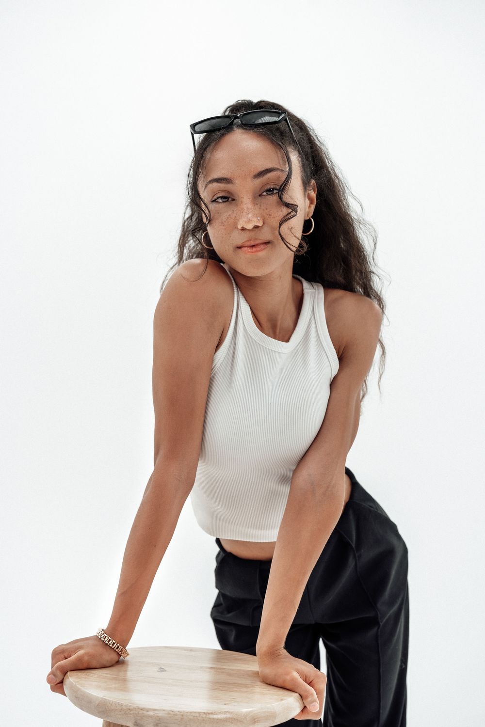 Young woman in white crop top and black pants leans on wooden stool, sunglasses on head, white background.