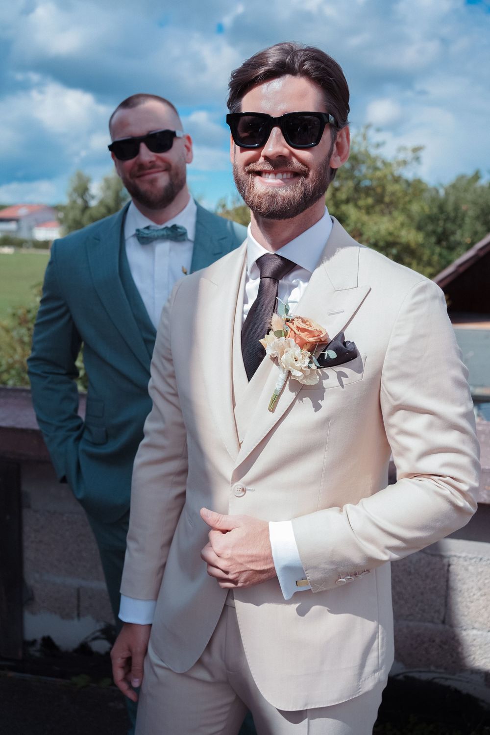A group of groomsmen in formal attire pose outdoors with some wearing teal suits and others in cream-colored suits.