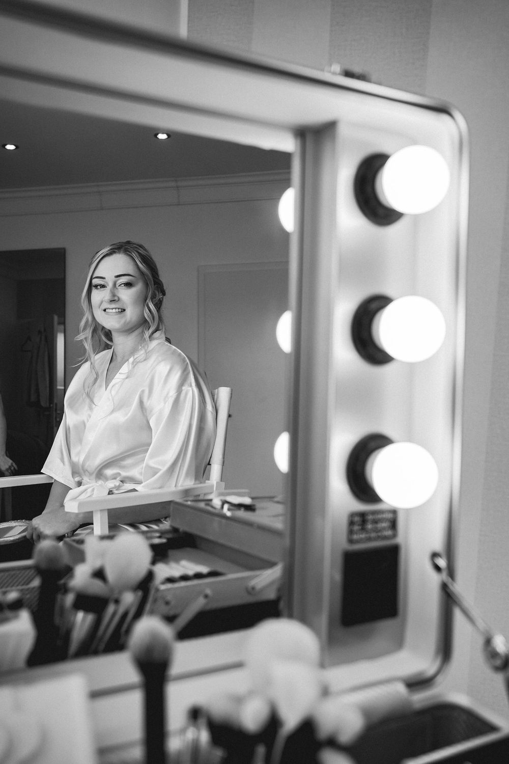 Black and white photo showing a makeup vanity with illuminated mirrors and someone sitting in a dressing room.