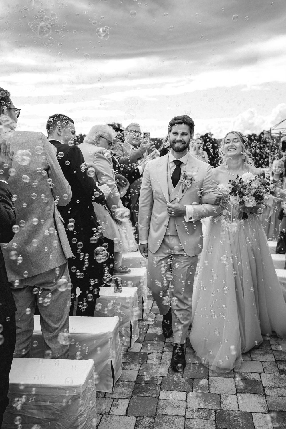 Wedding guests enjoy blowing bubbles during an outdoor celebration on a stone patio in black and white photography.