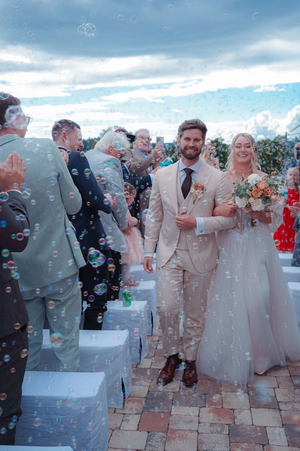 Wedding party celebrates on stone patio with bubbles floating through the air during outdoor reception celebration.