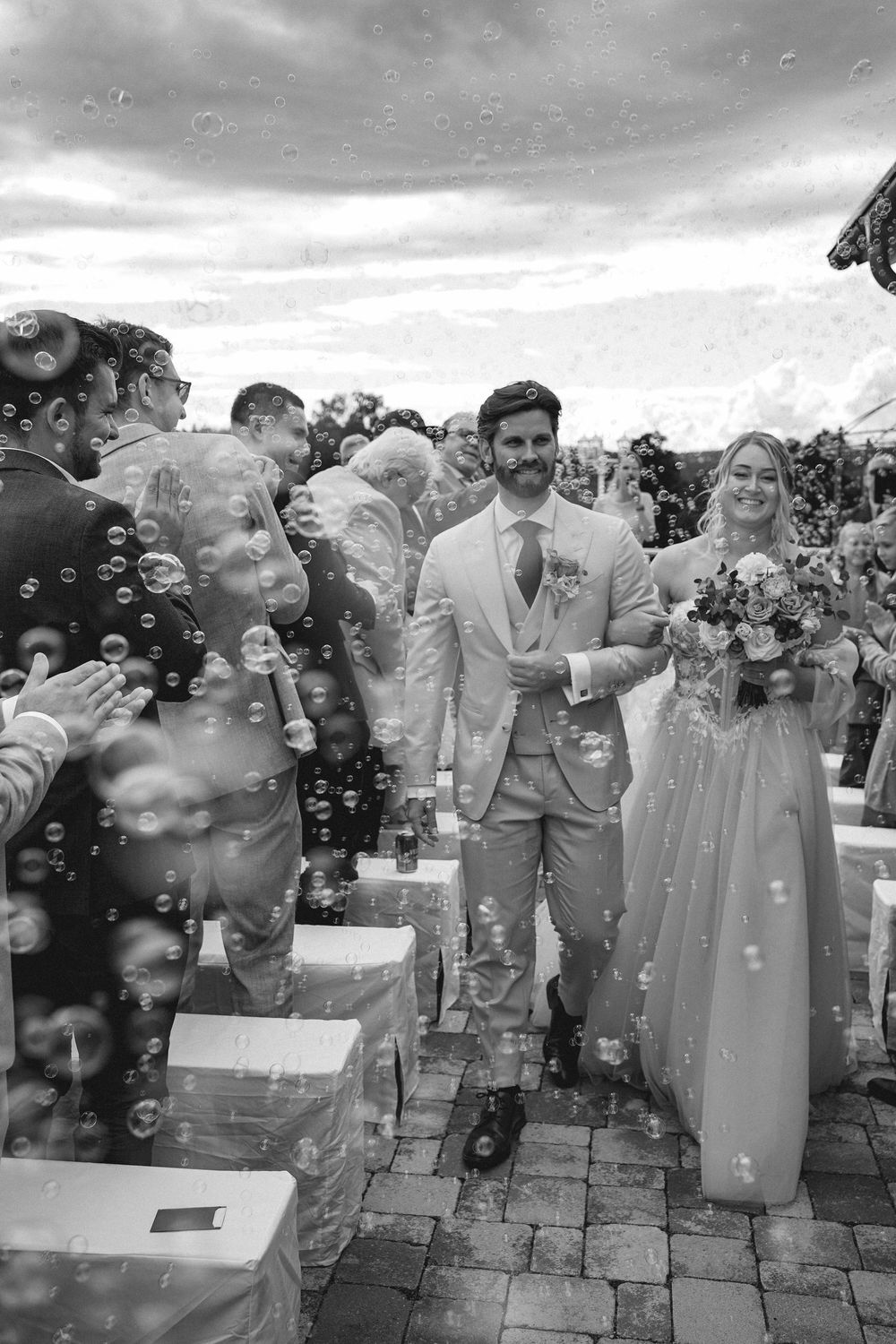 Wedding party members laugh and smile as soap bubbles float around them during an outdoor celebration on stone pavers.