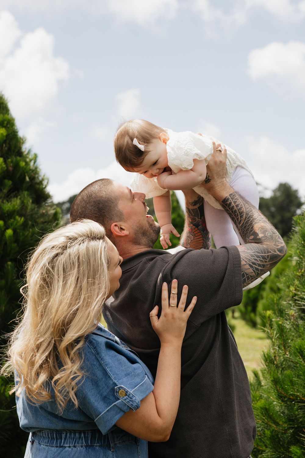 Happy family outdoors, father lifting baby girl while mother embraces them, surrounded by green trees.