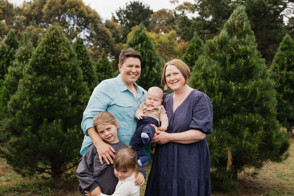 Family of four with newborn posing outdoors among lush green Christmas trees for a festive holiday portrait.