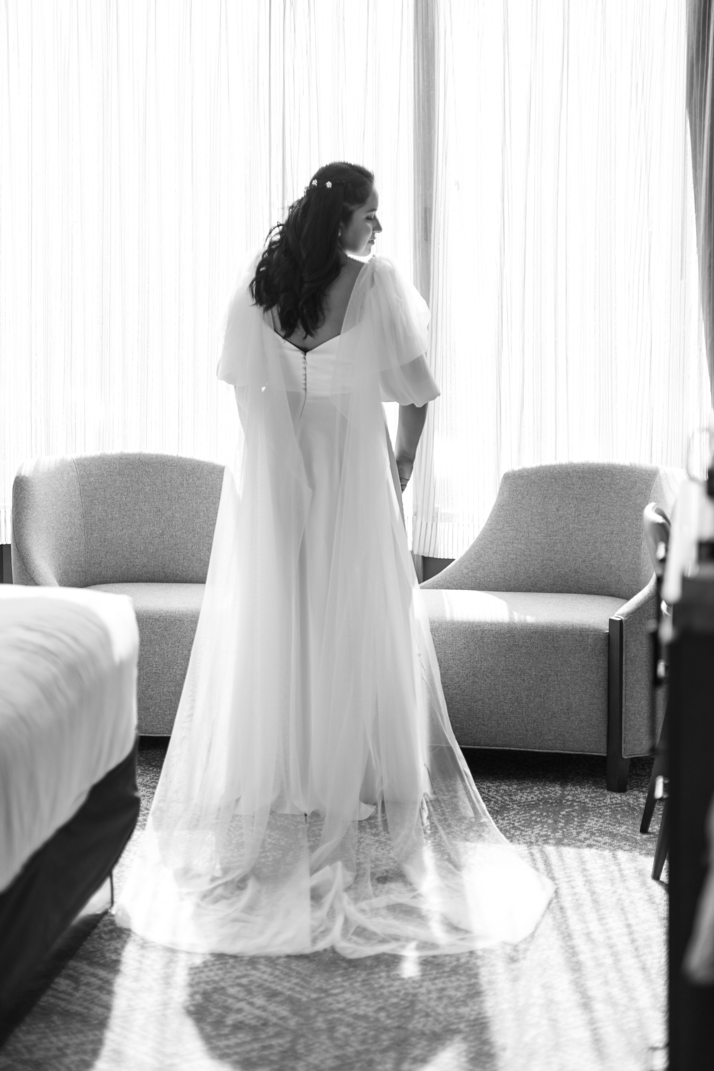 A bride in an elegant white wedding gown stands by a window in a contemplative pose.