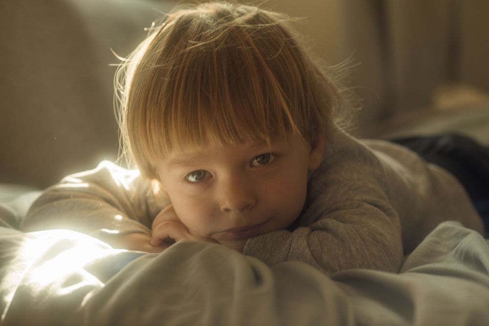 A young blonde toddler rests chin on hands, lying on white bedding in warm golden sunlight.