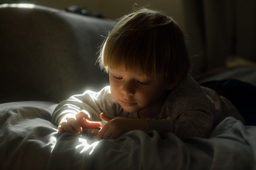 Young child lying on bed in soft dramatic lighting, looking at glowing phone screen in dark room.
