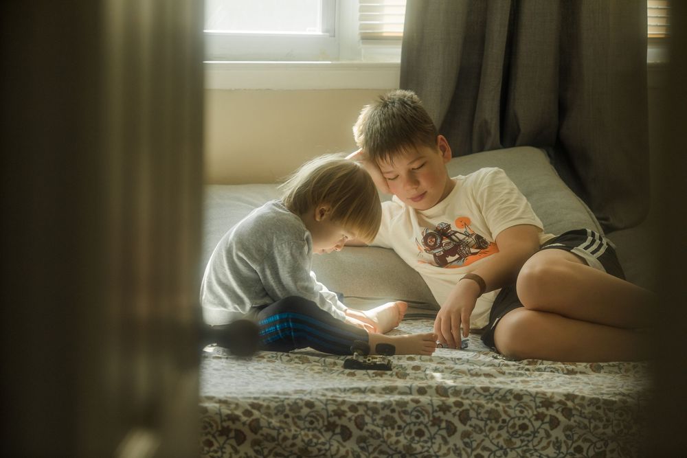 Two young boys sit on the floor together, engaged in a game, bathed in soft natural light near a window.