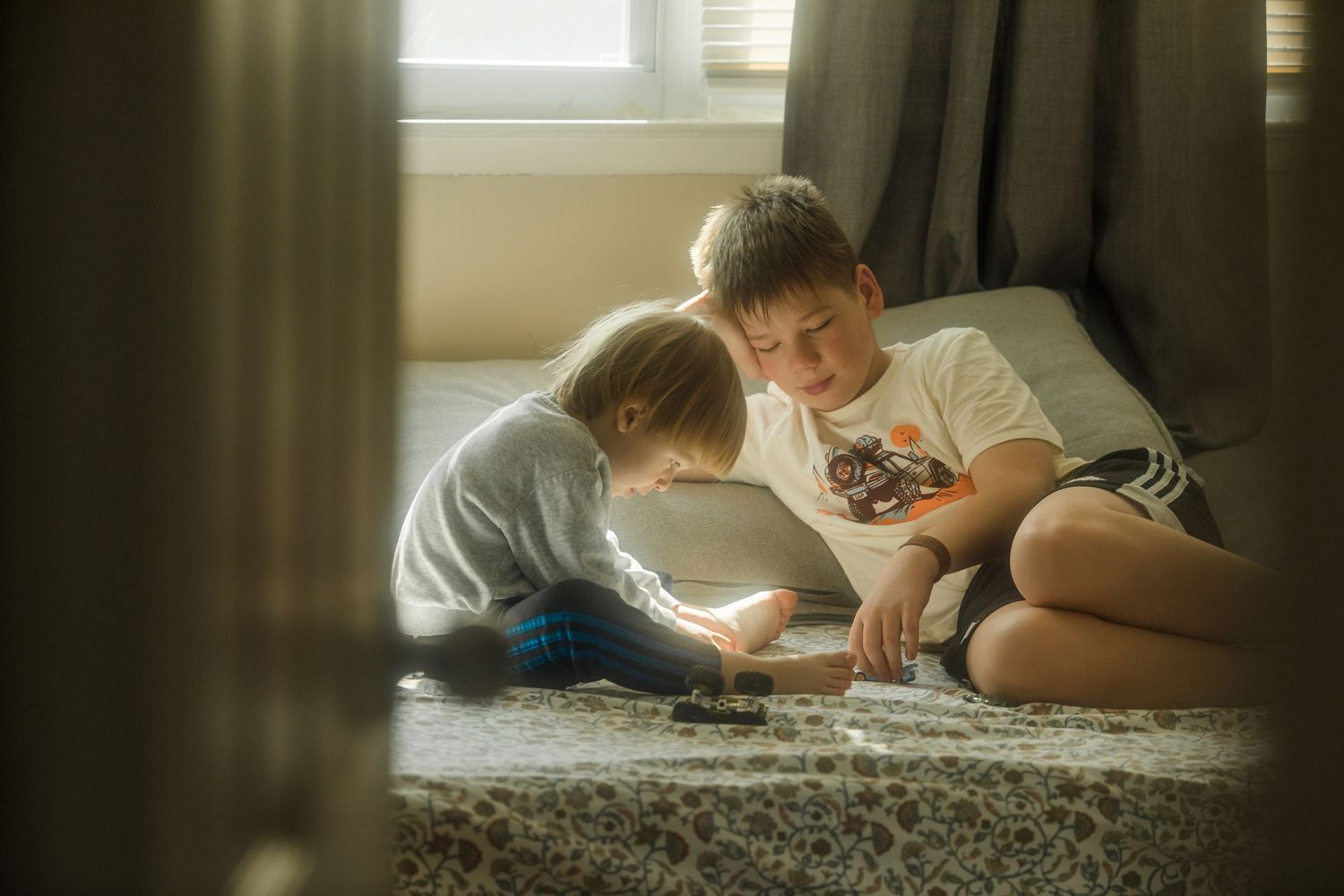 Two young boys sit on the floor together, engaged in a game, bathed in soft natural light near a window.