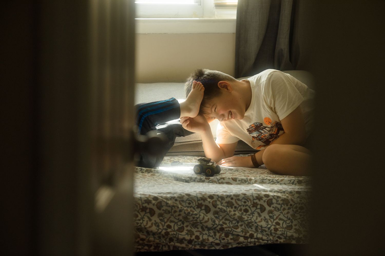 Young person sitting on bed in dimly lit room, leaning forward with head down in thoughtful or sad pose.