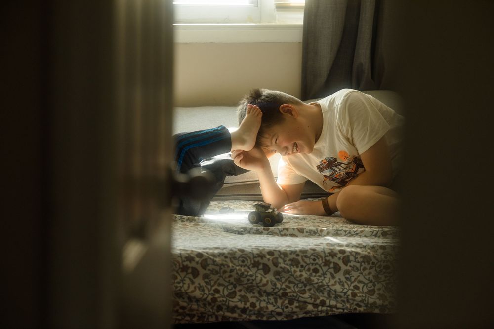 Young person sitting on bed in dimly lit room, leaning forward with head down in thoughtful or sad pose.