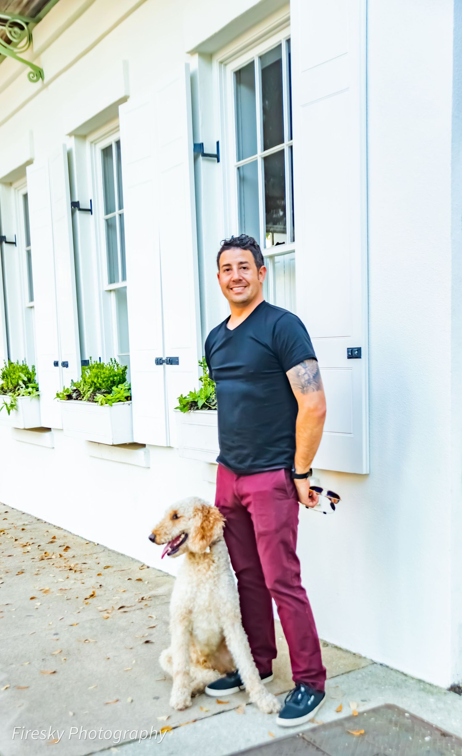 A casual portrait featuring a person in a navy shirt and burgundy pants standing with their golden doodle dog.