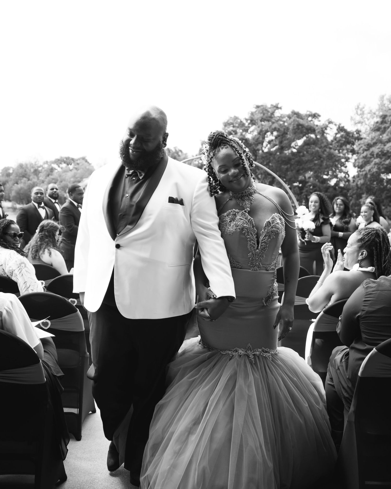 Black and white wedding photo of couple in formal attire walking through seated guests.