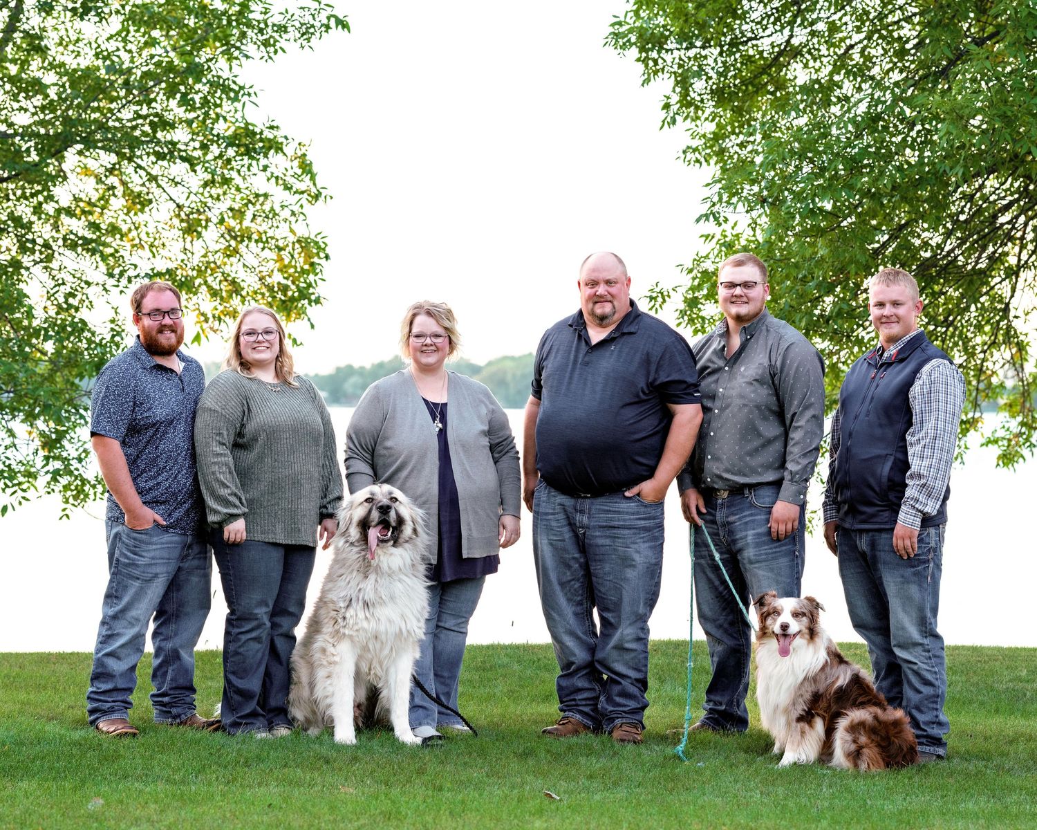 Extended family group of six with two dogs posing near a lakeside on a sunny day.