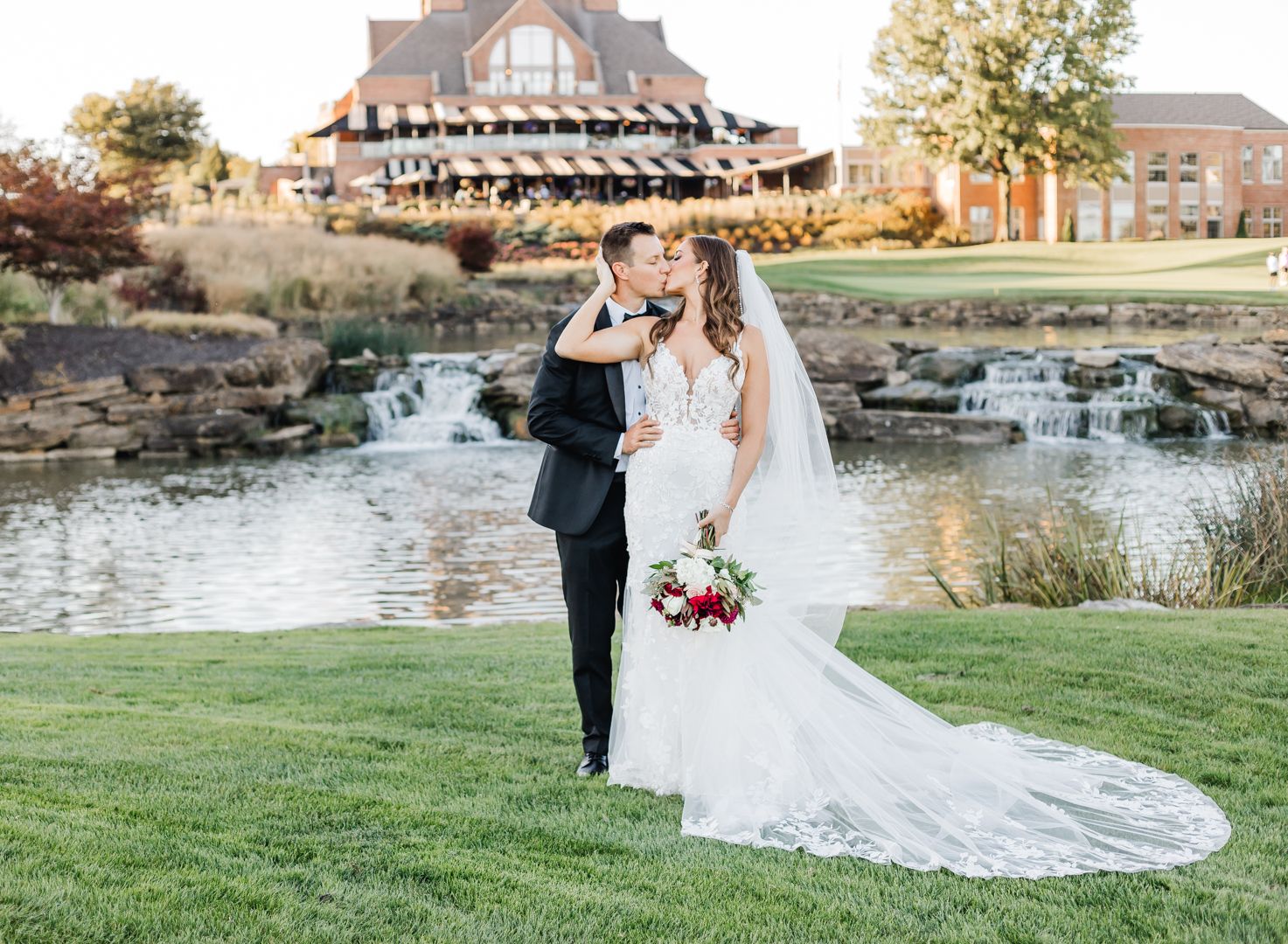 A couple poses together beside a waterfall in their wedding attire with a beautiful building in the background. Pittsburgh wedding Photog