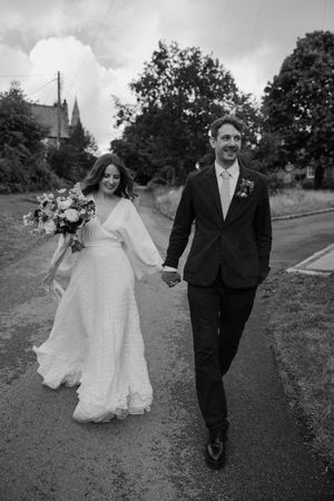 A bride in a flowing white dress and groom in a dark suit walk together on a country road with a church spire visible behind.