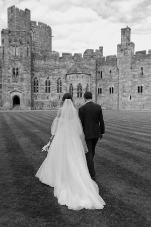 A bride and groom walk on the lawn towards Peckforton Castle on their wedding day.