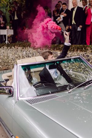 Groom in convertible car holding pink smoke flare during wedding as onlookers watch from behind.