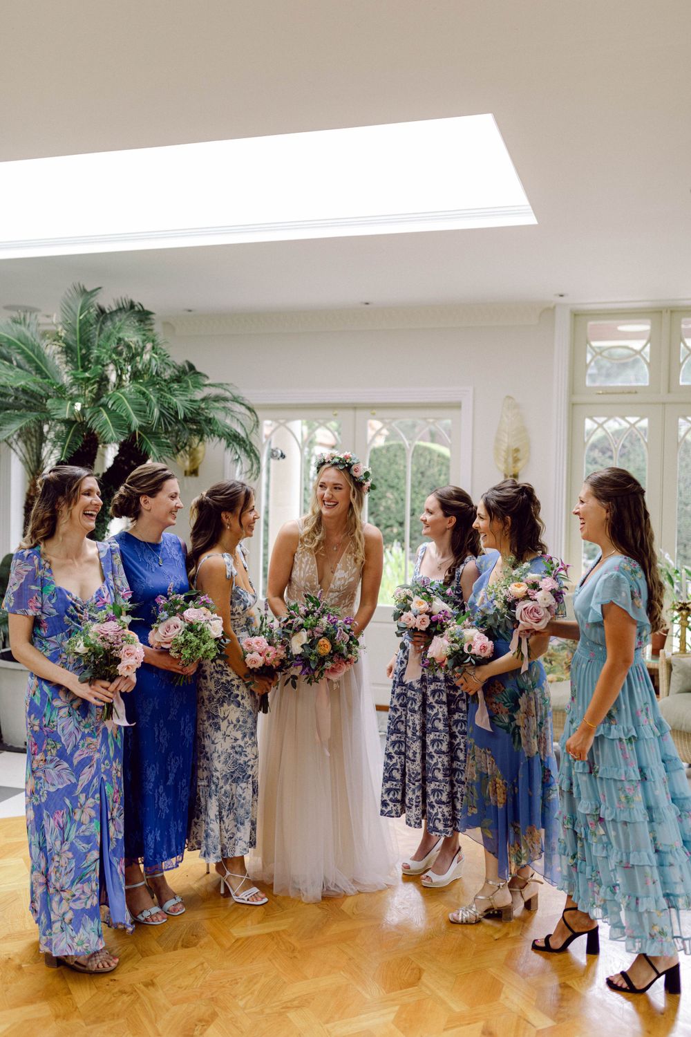 Wedding party in coordinated blue and floral dresses gather on a hardwood floor beneath a skylight in bright interior.