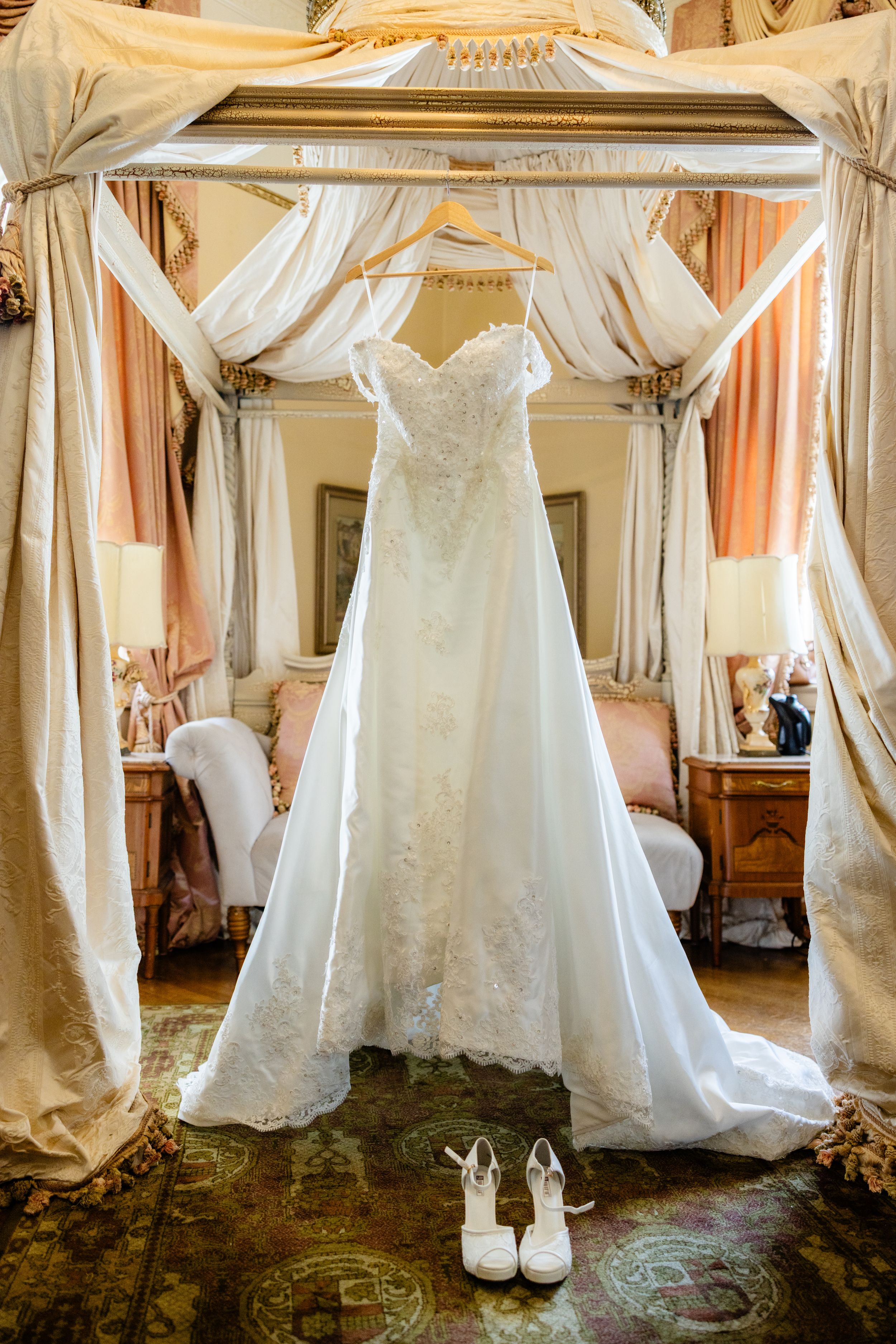 Elegant white lace wedding dress hanging on ornate stand with white heels beneath on vintage carpet.