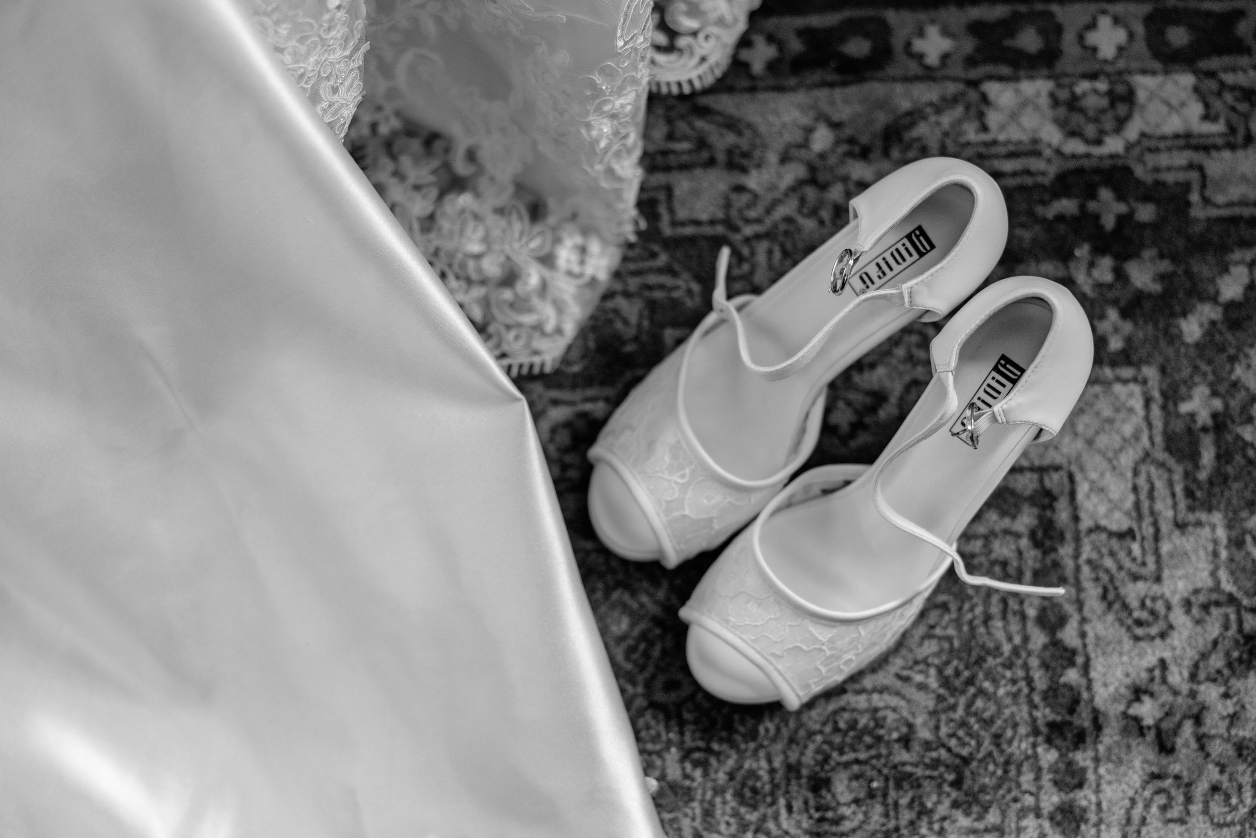 White wedding shoes placed on patterned carpet beside flowing fabric in monochrome.