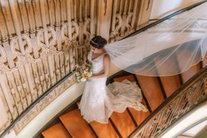 A bride in lace descends a curved staircase with flowing veil and bouquet.