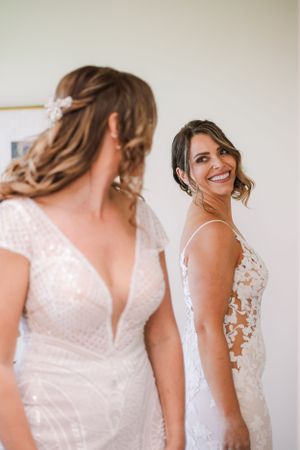 Two people in elegant white wedding dresses share a joyful moment while getting ready.