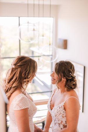 Two people in white lace wedding dresses share a tender moment near a bright window.