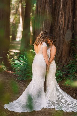 Two people in white wedding dresses embrace among tall redwood trees in a forest setting.