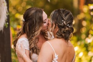Two people in delicate lace wedding dresses share an intimate kiss outdoors.