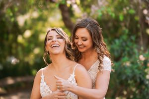 Two people in wedding dresses share a candid laughing moment in golden outdoor light.
