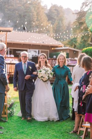Outdoor wedding processional with string lights and rustic venue in background.