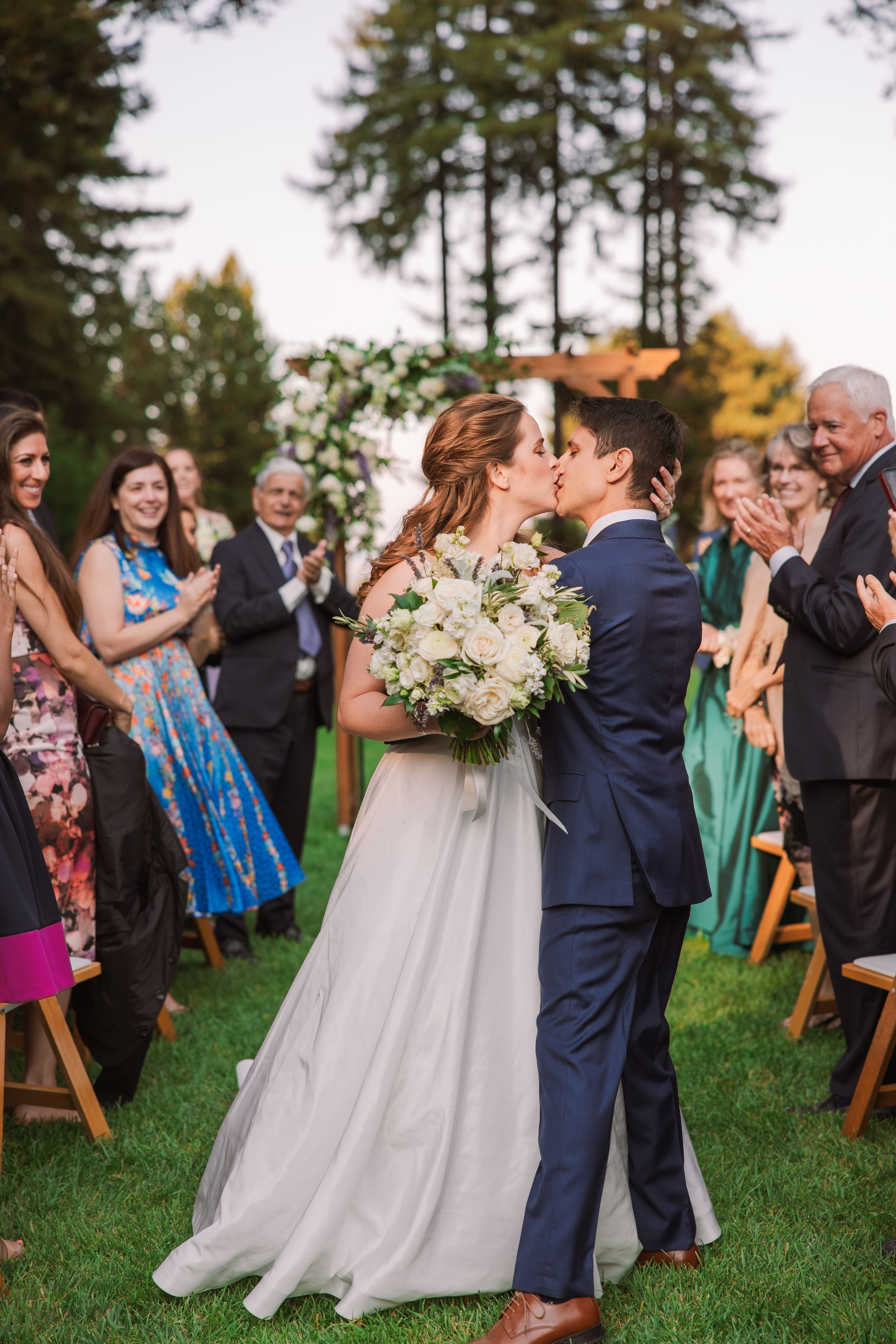 Newlyweds share their first kiss during outdoor ceremony as guests applaud.