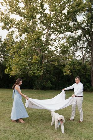 A couple in a grassy field holding a white sheet while their white dog stands between them on a sunny day.