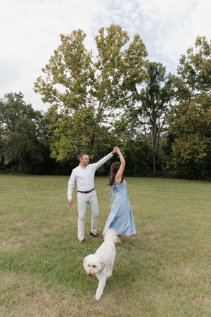 A couple dances playfully in a grassy field with their white dog on a sunny autumn day.