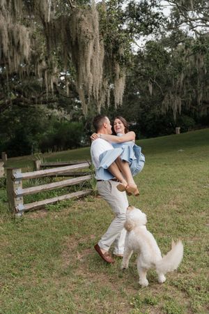 A couple dances playfully in a grassy field with a white fluffy dog while Spanish moss hangs from trees in the background.