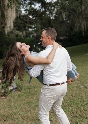 A romantic couple shares a joyful embrace while dancing outdoors in a park with Spanish moss hanging from trees.