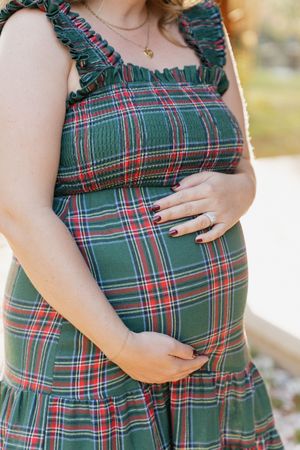 A pregnant figure in a green and red plaid dress with ruffled straps gently cradles their baby bump.