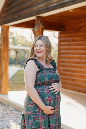 Woman wearing green plaid dress poses outdoors near rustic log cabin walls in natural lighting.