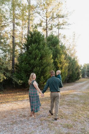 A couple walks hand in hand down a dirt path lined with evergreen trees on a sunny autumn day.