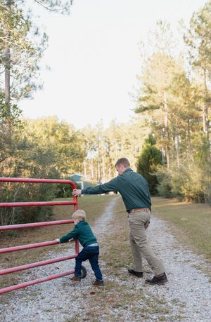 Two children playing and balancing on a red metal railing in a wooded outdoor area.
