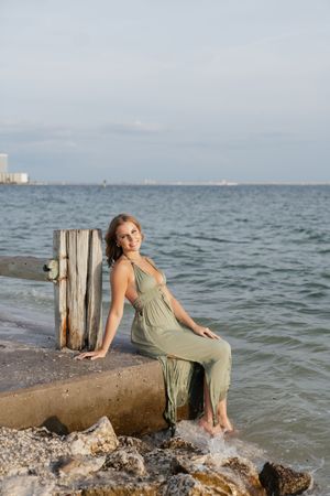 Young woman in olive green maxi dress sits on rocky coastal pier with ocean waves and city skyline in background.