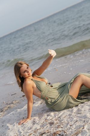 Woman in olive green dress posing on sandy beach with ocean waves in background.