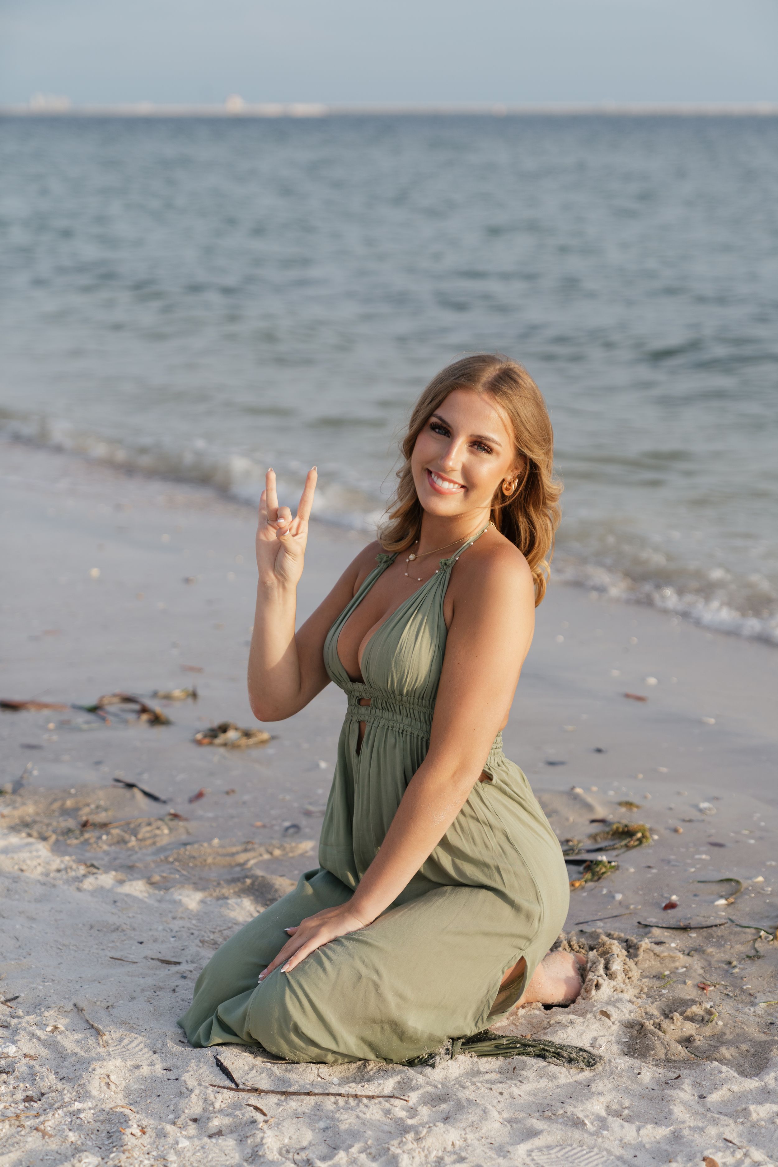Smiling blonde woman in green maxi dress sitting on sandy beach making peace sign with ocean in background.