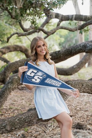 Smiling young woman holding a Georgia Southern University pennant while posing by a tree in a white dress.