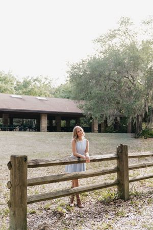 Woman in white dress leaning on rustic wooden fence at a scenic farm property with lush trees and pavilion.