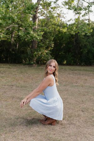Young woman in light blue dress and brown boots crouching gracefully outdoors on grass with lush green trees behind her.