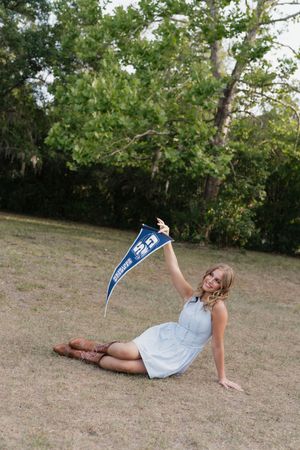 A young woman in a light blue dress and cowboy boots sits on grass, holding up a blue pennant flag outdoors.